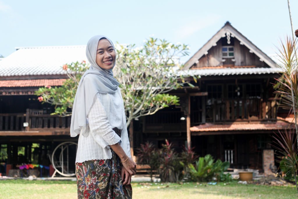 A young Malay Muslim woman wearing a hijab in front a traditional Malay house.